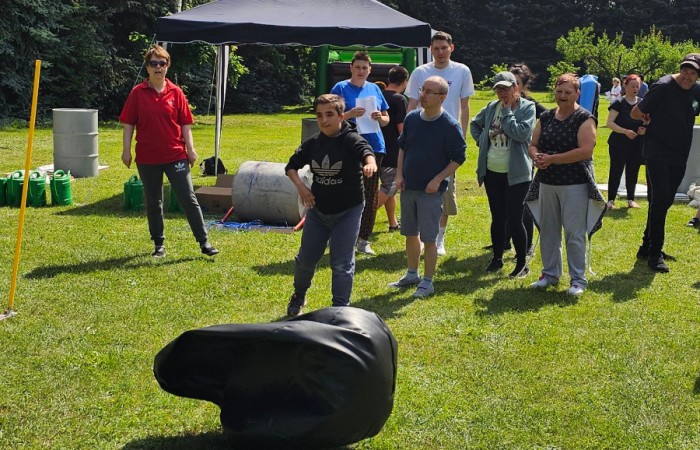 Kolejna osoba rzucająca poduszkę w kierunku wyznaczonego celu – Młoda kobieta w czarnej bluzie celuje do obiektu na trawniku, a wokół niej znajdują się inni uczestnicy.