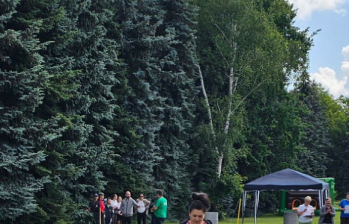 Kobieta uczestniczy w konkurencji sportowej, po trawie wzdłuż linii niesie dwie zielone ciężkie konewki. W tle widać sędzinę która mierzy czas w którym pokona dystans oraz innych zawodników dopingujących