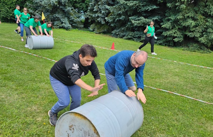 Konkurencja sportowa w której dwójka zawodników toczy po trawie starą szarą dużą beczkę, w tle widać zawodników innej drużyny którzy uczestniczą w tym samym wyzwaniu.