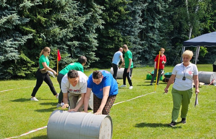 Konkurencja sportowa w której dwójka zawodników toczy po trawie starą szarą dużą beczkę, w tle widać zawodników innej drużyny którzy uczestniczą w tym samym wyzwaniu.