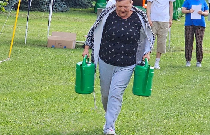 Kobieta uczestniczy w konkurencji sportowej, po trawie wzdłuż linii niesie dwie zielone ciężkie konewki. W tle widać sędzinę która mierzy czas w którym pokona dystans oraz innych zawodników dopingujących.