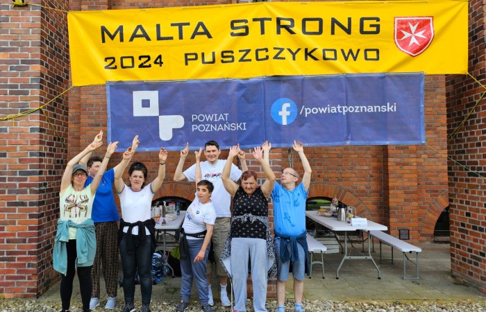 Grupa uczestników stojąca pod banerem „MALTA STRONG 2024 PUSZCZYKOWO” – Osoby w różnym wieku podnoszą ręce w geście radości, stojąc przed ceglaną budowlą. Za nimi wiszą banery z logo „Powiat Poznański” i odnośnikiem do mediów społecznościowych.