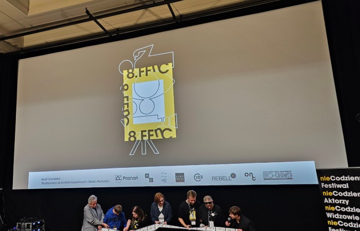 Zbliżenie na scenę, w tle wyświetlone logo 8 FFnC. Stoi na niej 7 osób z mikrofonami trzymających długą rolkę kamery.