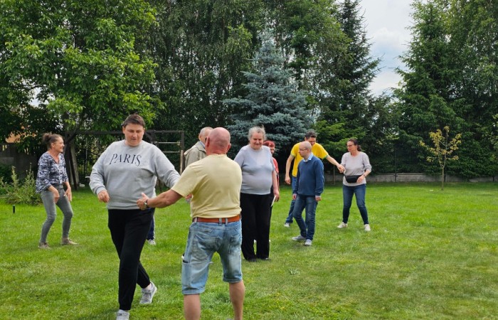 Grupa osób tańczy i bawi się na trawie, obok nich leży pies