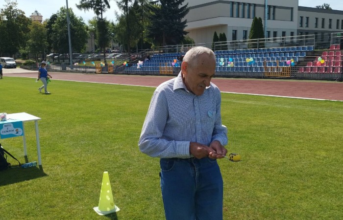 Starszy mężczyzna z łyżką i piłką w ręce przechodzi między żółtymi pachołkami na boisku.