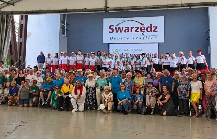 Duża grupa osób na wspólnym zdjęciu w hali, w tle napis „Swarzędz” i występujący artyści. 