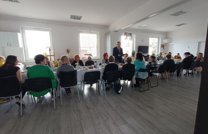 Przy stole nakrytym białym obrusem siedzi duża grupa ludzi, przed wspólnym posiłkiem przemawia mężczyzna ubrany w garnitur z czerwonym krawatem