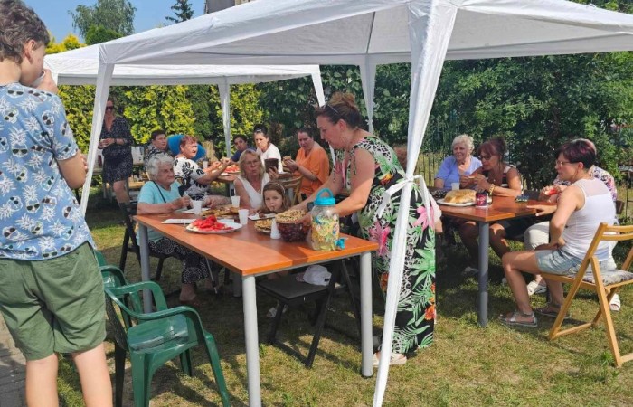 Grupa starszych i młodszych osób siedzi przy stołach pod białym namiotem w ogrodzie, rozmawiając i spożywając posiłki. 