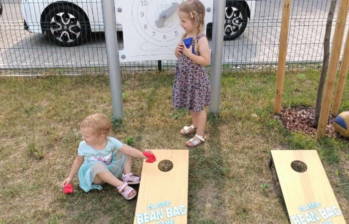 Dwie młode dziewczynki, jedna siedzi druga stoi na trawie, grają w grę Classic Bean Bag Game. W tle widać stojak z zabawką białego zegara, młode drzewo i białe auto.