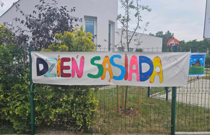 Na zielonym, siatkowym płocie wisi baner z kolorowym napisem Dzień Sąsiada na Działkowej 