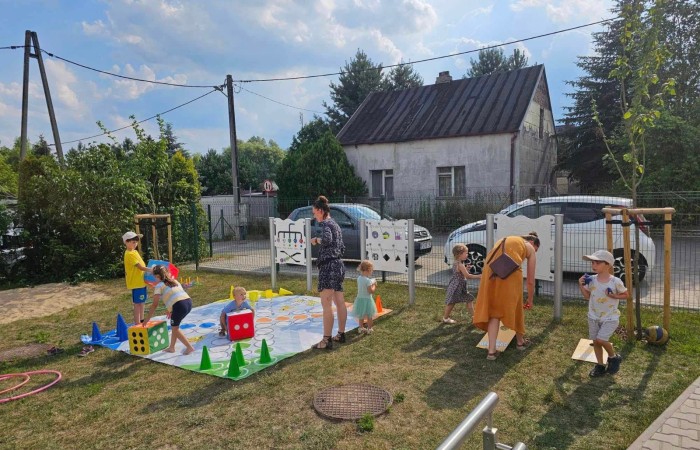 Grupa dzieci grających w gry plenerowe, rzucających woreczki do celu i używających dużych kostek do gry. W tle ogrodzenie i domy.