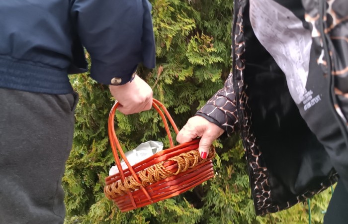 Mężczyzna i kobieta stoją blisko krzewów, trzymając koszyk wypełniony foliowymi torebkami z przedmiotami. Ręce obojga są w ruchu, jakby coś w koszyku układali lub przeszukiwali