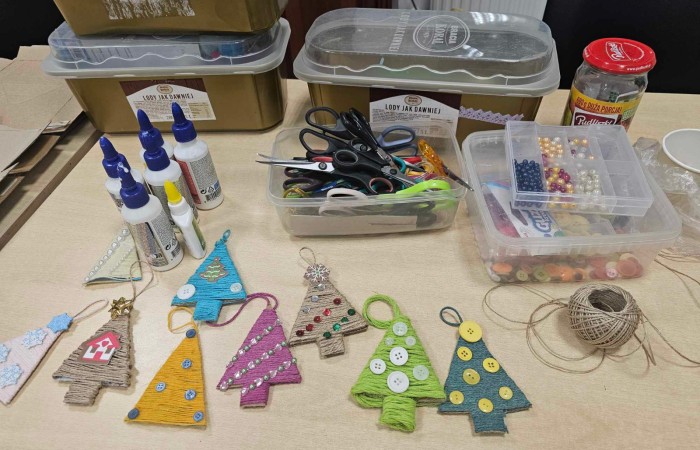 Własnoręcznie robione zawieszki na choinkę w jej kształcie w różnych kolorach i z rożnymi dodatkami np. guzikami. W tle materiały do robótek ręcznych jak nożyczki, sznurki, perełki, kleje.