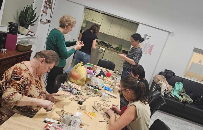 Grupa kobiet własnoręcznie robi zawieszki w kształcie choinek. Na stole leży wiele materiałów do pracy.