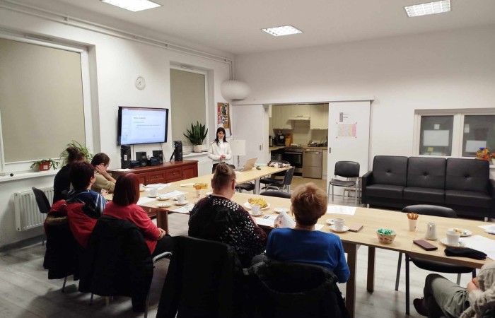 Grupa osób siedzi przy długim stole, z filiżankami i talerzykami. Słuchają prezentacji wyświetlanej na telewizorze przez kobietę w białym swetrze.