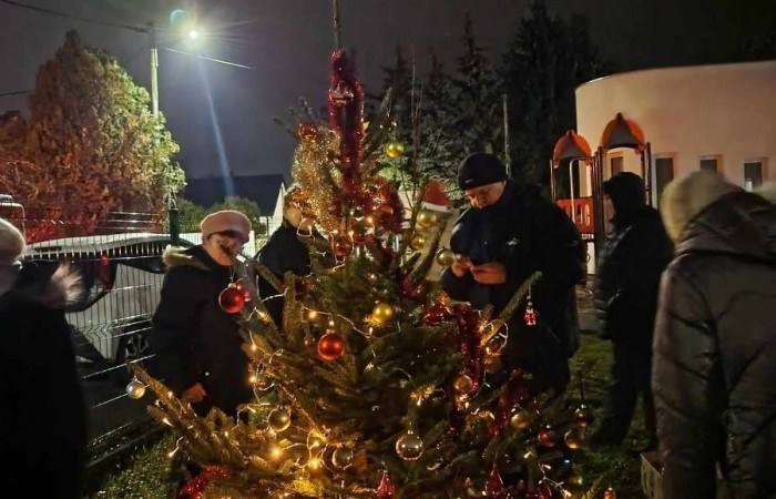 Kilka osób stojących wokół pięknie oświetlonej choinki na zewnątrz, zdjęcie wykonane wieczorem przy sztucznym świetle.