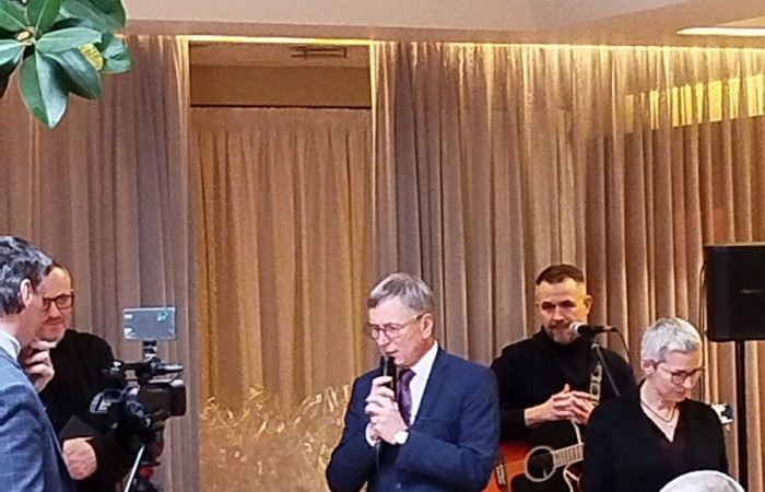 Dwie uśmiechnięte kobiety siedzące przy stole podczas imprezy, w tle widać grupę osób tańczących przy akompaniamencie gitarzysty