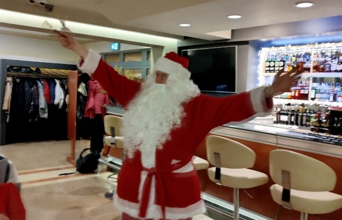 Osoba przebrana za Świętego Mikołaja stoi z rozłożonymi rękami w sali, w tle bar i elementy wyposażenia wnętrza