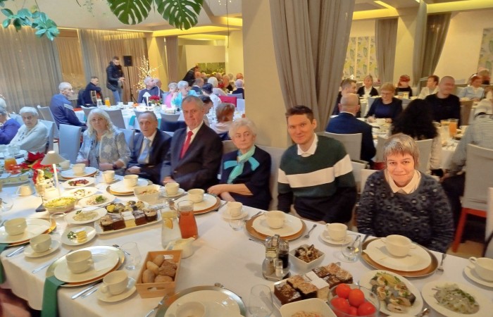 Stół zastawiony talerzami pełnymi przekąsek i ciastek. W tle widoczne osoby siedzące przy drugim stole