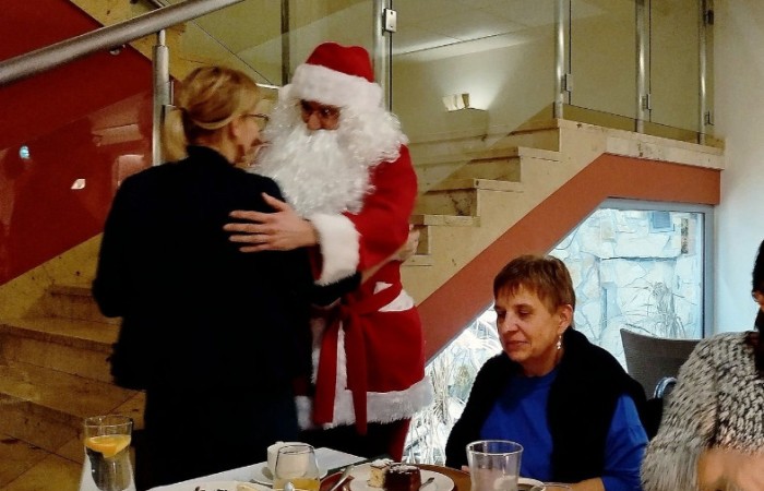 Osoba przebrana za Świętego Mikołaja wręcza prezent kobiecie, w tle widoczne są inne osoby oraz schody prowadzące na piętro