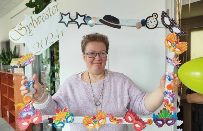 Uśmiechnięta osoba pozująca do zdjęcia w kolorowej, ozdobnej ramce z napisem 'Sylwester' i różnymi zabawnymi dekoracjami.