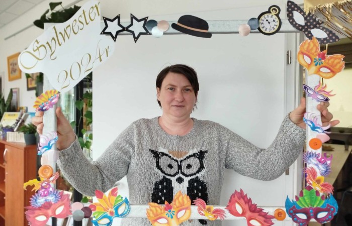 Uśmiechnięta osoba pozująca do zdjęcia w kolorowej, ozdobnej ramce z napisem 'Sylwester' i różnymi zabawnymi dekoracjami.