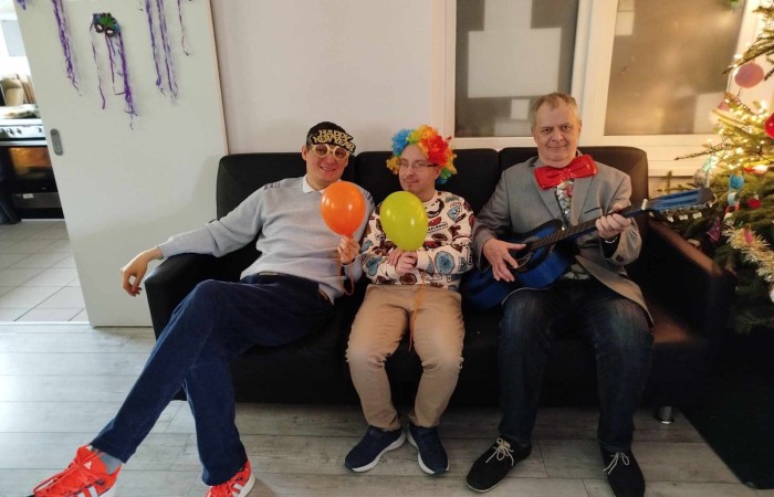 Dwie kobiety siedzące przy stole, ubrane w zabawne akcesoria, w tle tańczące osoby oraz mężczyzna grający na gitarze