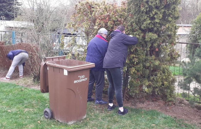 Trzy osoby wykonujące prace ogrodowe przy żywopłocie. Obok stoi brązowy pojemnik na odpady biodegradowalne oznaczony napisem „BIOODPADY”.