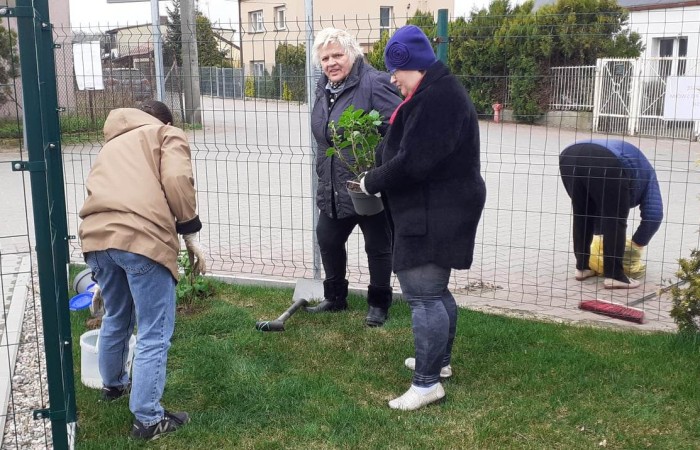 Grupa osób pracuje przy ogrodzeniu. Jedna osoba podlewa roślinę, druga trzyma doniczkę z sadzonką, trzecia obserwuje, a czwarta zamiata chodnik po drugiej stronie ogrodzenia. W tle widać zabudowania i zaparkowane samochody.