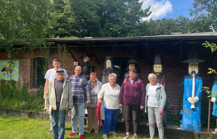 Uczestnicy ŚDS pozują do zdjęcia na zewnątrz muzeum pszczelarstwa. Za nimi stoi 6 męskich figur wyrzeźbionych w drewnie.